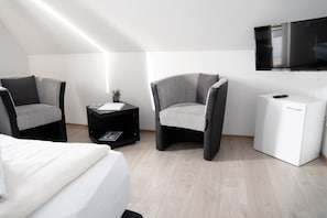 Habitació moderna amb llit, dos seients, taula petita amb una planta, moble blanc i televisió de pantalla plana penjada a la paret.