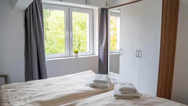 A bedroom with a bed, a window with curtains, a small plant on the windowsill, and a mirror on the wall.