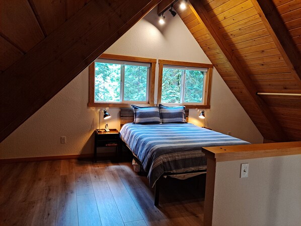 A bedroom with a sloped ceiling, wooden beams, a bed with striped bedding, two bedside lamps, and a window with a view of trees. - Mt cabin in private community close to Mt Baker (Deming)