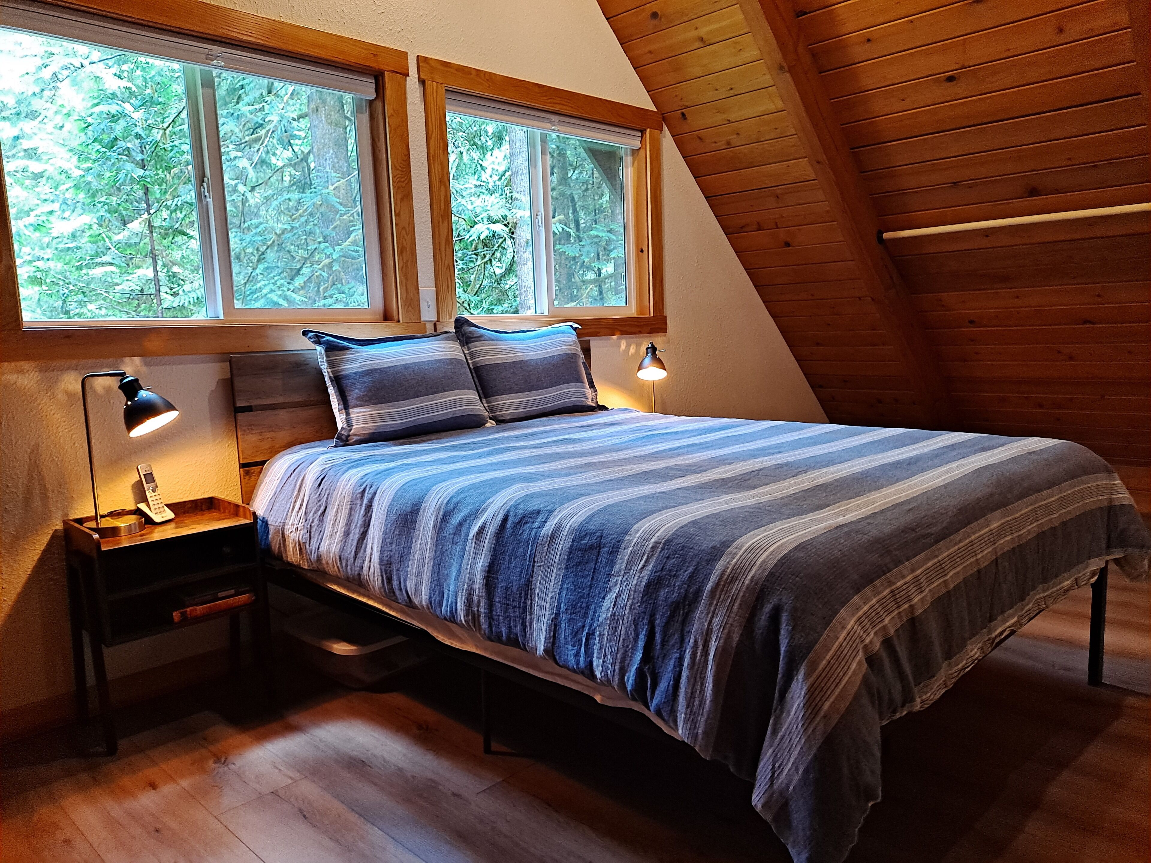 A bedroom with a sloped ceiling, wooden walls, a bed with striped bedding, two bedside lamps, and a nightstand with a remote control.
