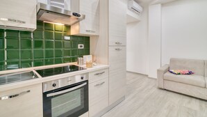 A modern kitchen with a built-in stove, oven, and stainless steel range hood. There is a green tiled backsplash and a beige sofa in the adjacent living area. - Eclisse House Few Steps From The Sea, Augusta, Italy (Augusta)
