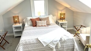 A bedroom with a sloped ceiling, a large bed with patterned bedding, two bedside tables with lamps, a chair, and a window. - Beach House ∆ Prime Location ∆ Steps to the Ocean (Old Orchard Beach)