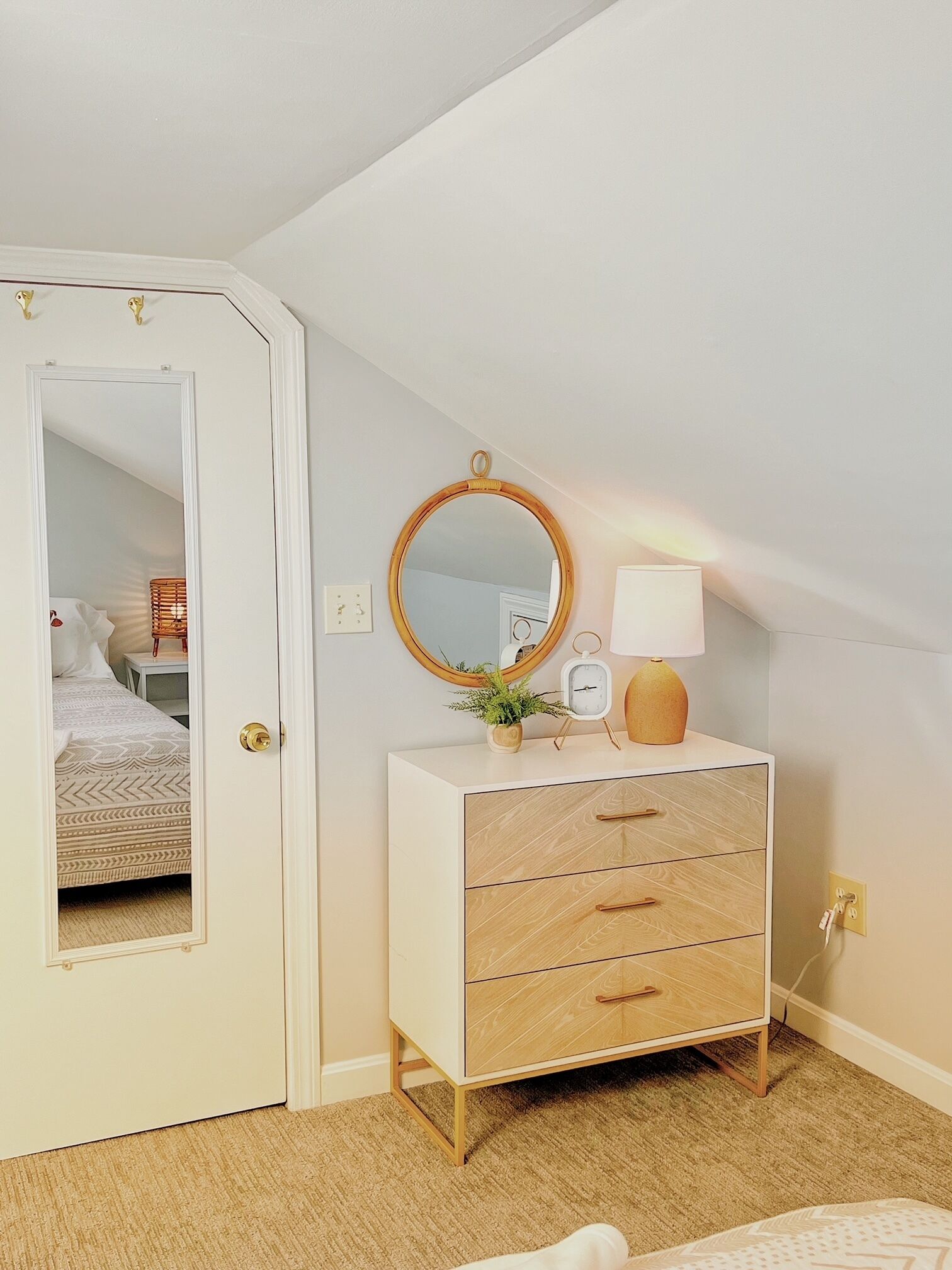 Een slaapkamer met een witte kapstok, een ronde spiegel, een bedlampje en een deur die naar een andere kamer leidt.