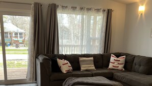 A living room with a sectional sofa, a patterned rug, and a view of a house through a window with curtains. - 🇨🇦 Lake Winnipeg Fishing All-Seasonal Retreat (Winnipeg Beach)