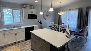 A modern kitchen with white cabinets, a central island, pendant lights, and a dining area with a table and chairs. - 🇨🇦 Lake Winnipeg Fishing All-Seasonal Retreat (Winnipeg Beach)