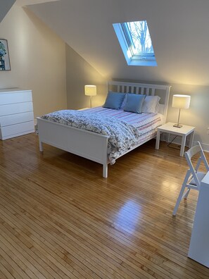A bedroom with a skylight, a bed with floral bedding, two bedside tables with lamps, a white dresser, and a wooden floor. - Cheerful 5 bed Residential house. (Bridgeport)