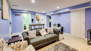 Ein Wohnzimmer mit grauem Sofa, einem Couchtisch und einem Teppich. Eine Tür mit grünem Rahmen und eine Wanduhr sind ebenfalls zu sehen.