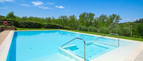 Una piscina rectangular con agua azul cristalina, rodeada por una terraza pavimentada y una zona verde con árboles.