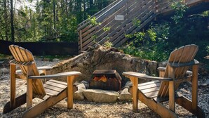 Deux fauteuils Adirondack sont placés face à un foyer, avec une échelle en bois à proximité, dans une zone boisée.