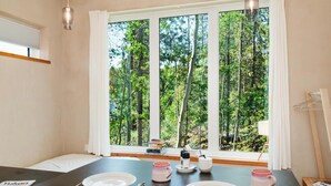 Un coin repas avec une table dressée pour deux personnes, une fenêtre donnant sur la forêt et une petite étagère avec des livres et une bouilloire.