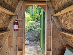 A wooden attic room with a green door leading outside, a fire extinguisher on the wall, and a view of greenery outside. - A-Frame Shelter (T15)  - Oceanside Views (Port Angeles)