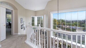 A spacious hallway with a white railing, large windows offering a view of the neighborhood, and framed pictures on the walls.