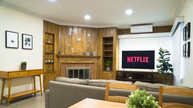 Ein Wohnzimmer mit einer Holz-Wandleuchte, einem Kamin, einem Fernseher, der 'NETFLIX' zeigt, und einem Essbereich mit Holztisch und Stühlen.