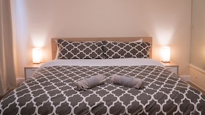 Ein ordentlich bezogenes Bett mit einem gemusterten Überwurf, zwei Nachttischlampen und einem zusammengerollten Handtuch.