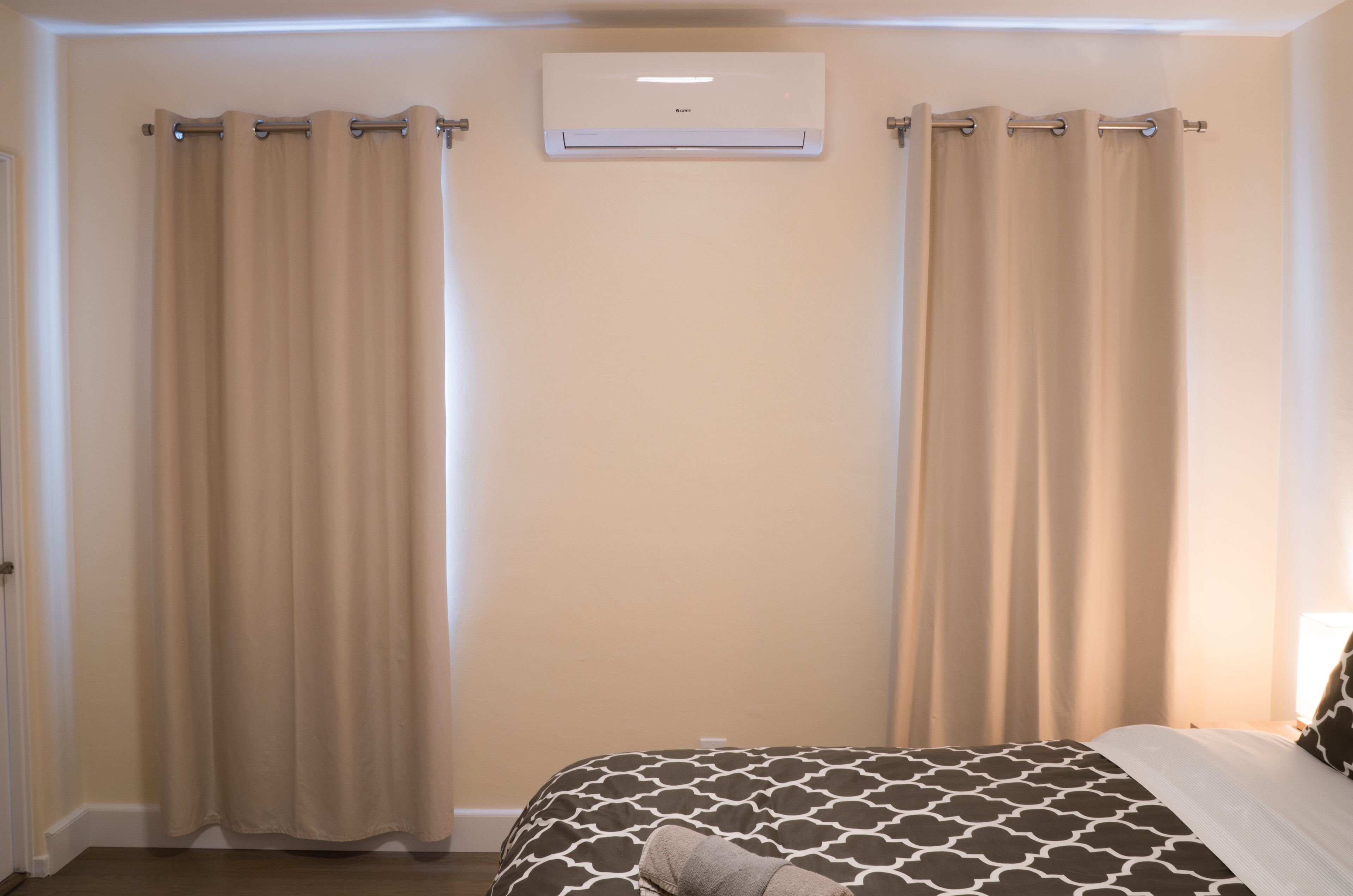 A bedroom with beige curtains, a patterned bedspread, and a wall-mounted air conditioner.