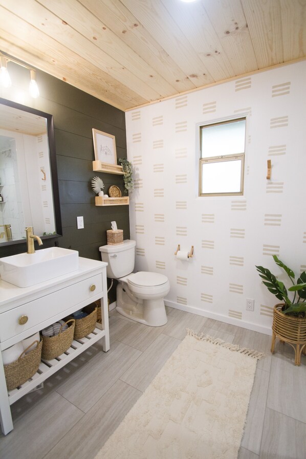 A bathroom with a white sink, toilet, and vanity. There is a mirror above the sink, a window above the toilet, and a plant on the floor.
