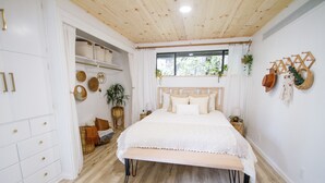 Un dormitorio con techo de madera, una cama con ropa de cama blanca, una ventana con cortinas y una pared decorada con objetos.