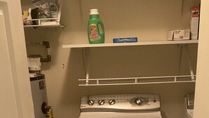 Ein kleiner Waschraum mit Waschmaschine und Trockner, einem Warmwasserbereiter und Regalen mit Reinigungsmitteln.