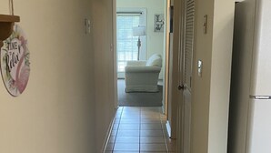 Ein schmaler Korridor mit Fliesenboden, einem Kühlschrank auf der rechten Seite und einer Tür zu einem Raum mit Fenster und einer Couch.