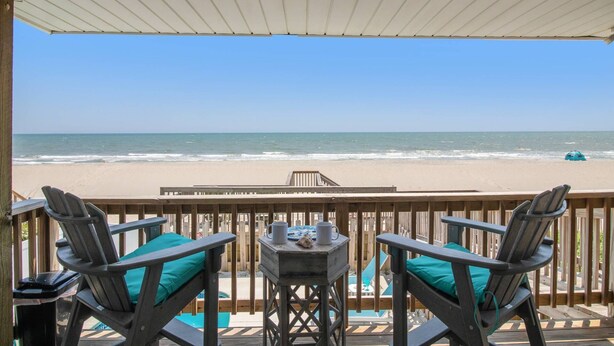 Une terrasse en bois avec deux chaises et une petite table, offrant une vue dégagée sur la plage et l’océan.