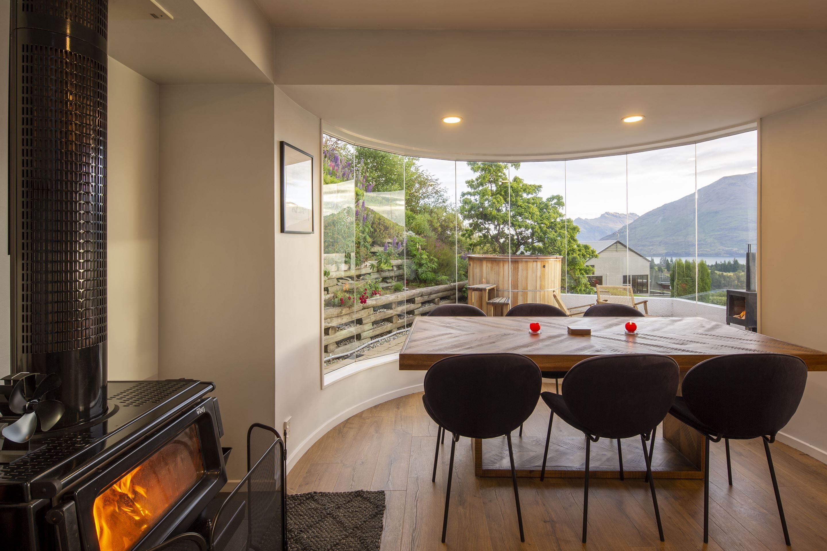 A modern dining area with a large table and four chairs, a wood-burning stove, and a large window offering a scenic mountain view.