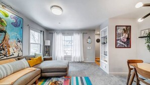 Een moderne woonkamer met een bank, een kleurrijk vloerkleed, een salontafel, en een eethoek met een houten tafel en stoelen.