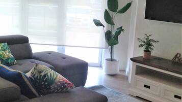 Una sala de estar moderna con un sofá gris, una mesa de centro, un televisor en la pared y una planta en maceta.