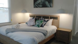 Une chambre à coucher avec un lit en bois, du linge de lit blanc, une grande fenêtre et une œuvre d’art accrochée au mur.