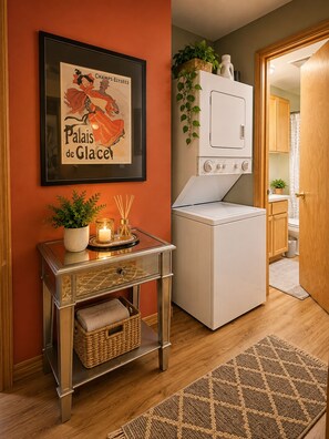 Un espace buanderie avec un lave-linge et un sèche-linge, une affiche encadrée au mur, une petite table avec une bougie et un panier.