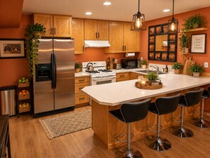 A modern kitchen with wooden cabinets, stainless steel appliances, a central island with bar stools, and decorative plants. - Charming & Cozy Apartment w/ firepit & patio. (Kenmore)