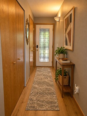 Un couloir avec une porte en bois, une porte vitrée donnant sur l’extérieur, un tapis, une lampe fixée au mur et une petite table sur laquelle se trouvent une plante et un bol.