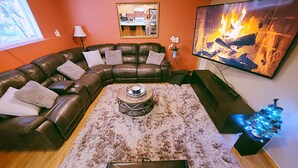 A living room with a sectional sofa, a coffee table, a rug, a flat-screen TV, and a fireplace painting. - Charming & Cozy Apartment w/ Hot tub patio. (Kenmore)