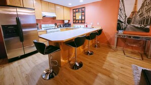 A modern kitchen with wooden cabinets, a stainless steel refrigerator, a gas stove, and a large island with bar stools. - Charming & Cozy Apartment w/ Hot tub patio. (Kenmore)