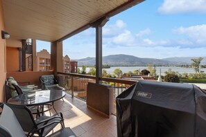 Un balcón con una mesa y sillas de vidrio, una parrilla y vistas a un lago y montañas.