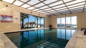 Une piscine intérieure avec des parois en verre transparent, un panneau indiquant la présence d’un sauveteur et une vue sur le port de plaisance.