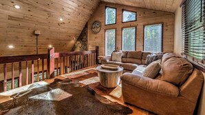 A cozy cabin interior with a wooden ceiling, a sofa, a coffee table, and a large window with blinds. - The Edison - Hot Tub | Shuffleboard | Arcade Game | Firepit (Blue Ridge)