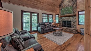 A spacious living room with a stone fireplace, a large window with a football game, and a cozy seating area. - The Edison - Hot Tub | Shuffleboard | Arcade Game | Firepit (Blue Ridge)