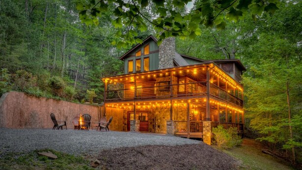 A two-story log cabin with a stone chimney, surrounded by a forest. - The Edison - Hot Tub | Shuffleboard | Arcade Game | Firepit (Blue Ridge)