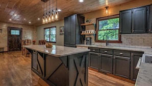 A spacious kitchen with a large island, dark cabinetry, and a window with a view of greenery. - The Edison - Hot Tub | Shuffleboard | Arcade Game | Firepit (Blue Ridge)