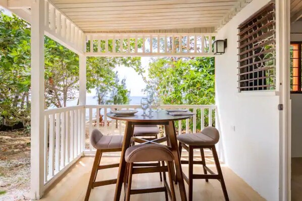 A covered outdoor seating area with a table and chairs, surrounded by white railings and greenery.