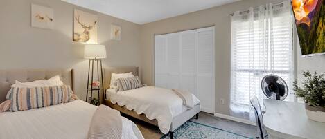 A bedroom with two beds, a desk, a chair, a lamp, a fan, and a window with curtains.