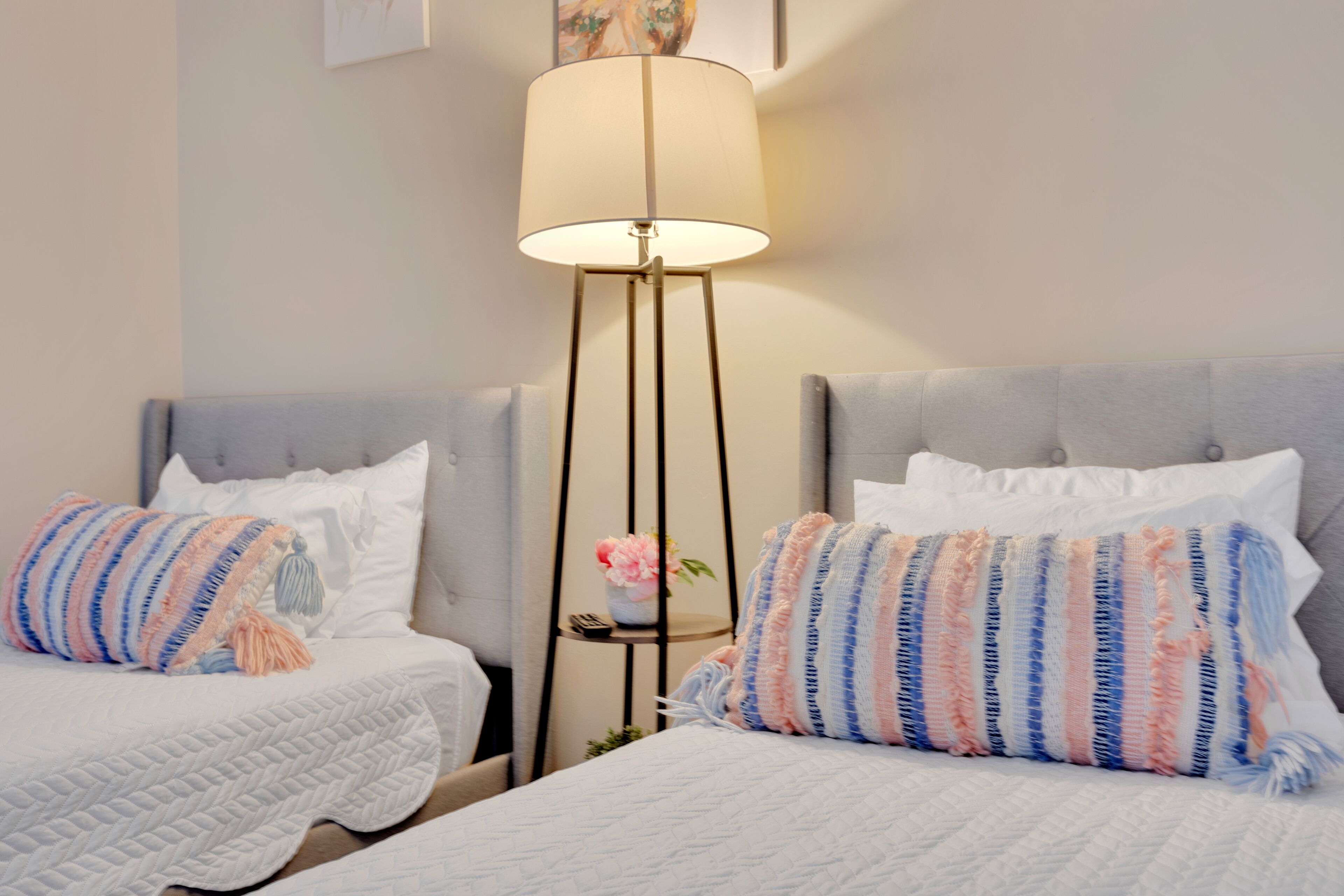 Ett hotellrum med två sängar, var och en med ett vitt, vadderat sängöverkast och randiga kuddar. Mellan sängarna står ett litet bord med en golvlampa med en beige lampskärm.