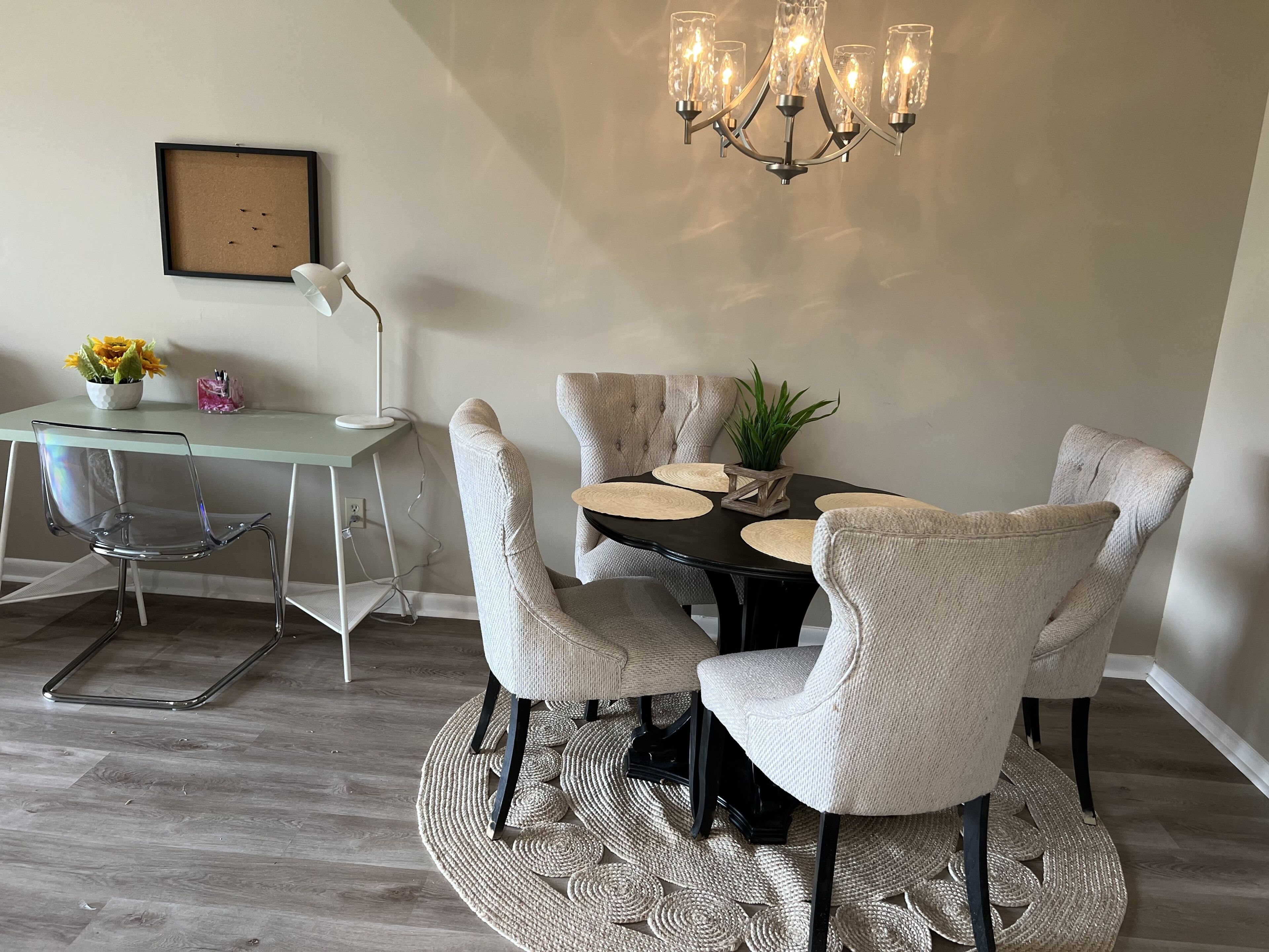 Een eetruimte met een ronde tafel, zes stoelen, een kroonluchter, een ingelijst schilderij aan de muur, een bureau met een computer en een plant in een pot.