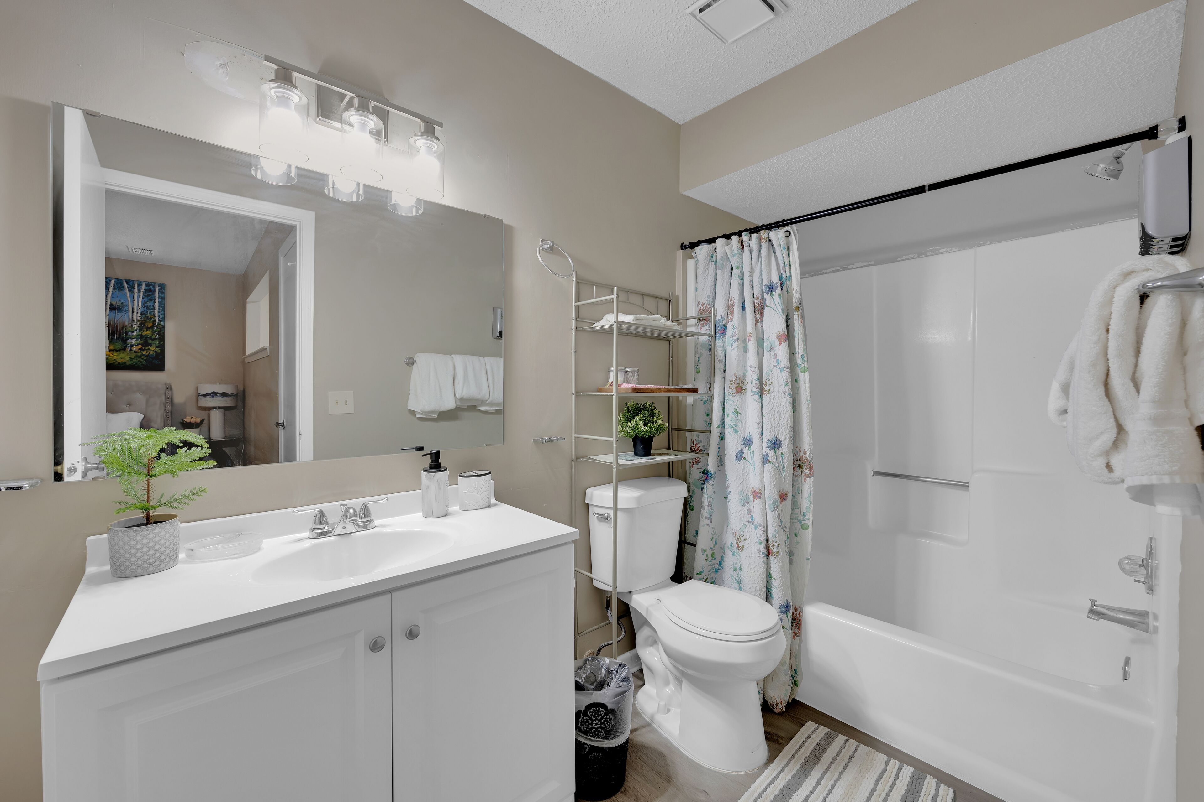 A modern bathroom with a white sink, toilet, and bathtub. There is a mirror above the sink, a shower curtain, and a potted plant on the sink.