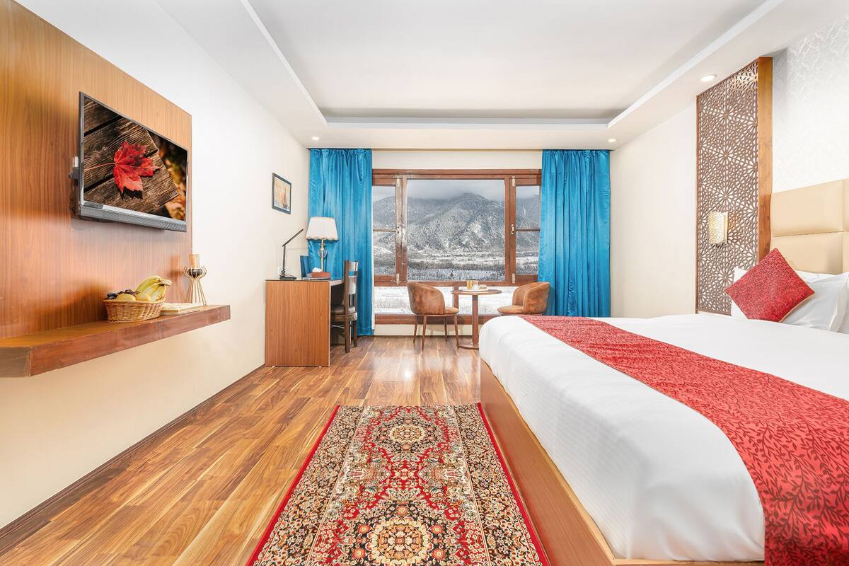 Habitación de hotel con una cama grande, un escritorio con silla, una mesita con dos sillas, un televisor montado en la pared y una ventana con vista a la montaña.