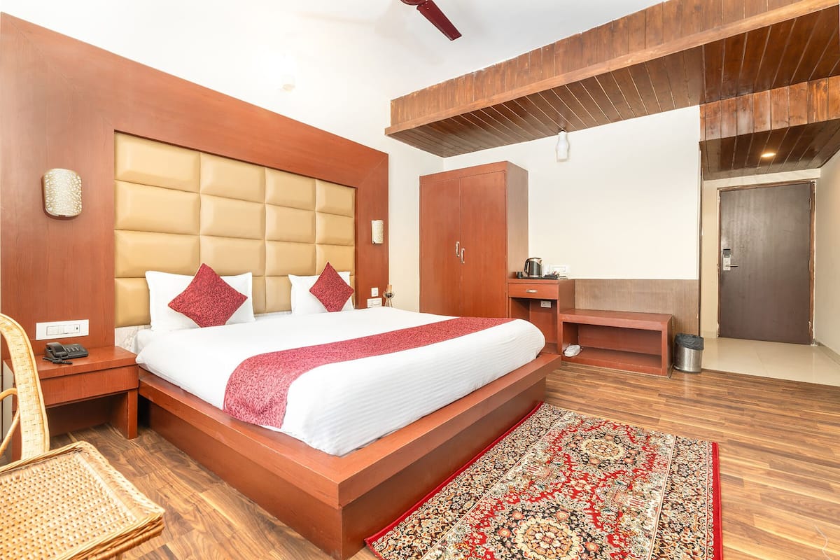 Una habitación de hotel con una cama grande, cabecera de madera, un pequeño escritorio, un armario, un cubo de basura y una silla de mimbre.