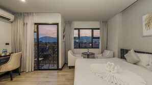 A hotel room with a large bed, a desk, a chair, a small table, and a balcony with a view of mountains and a cityscape. - Coco Paradiso Phuket Hotel (Chalong)
