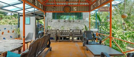 A covered outdoor seating area with wooden benches and tables, surrounded by a metal frame and glass roof, with a clock on the wall and a sign reading "Sauna Herb".