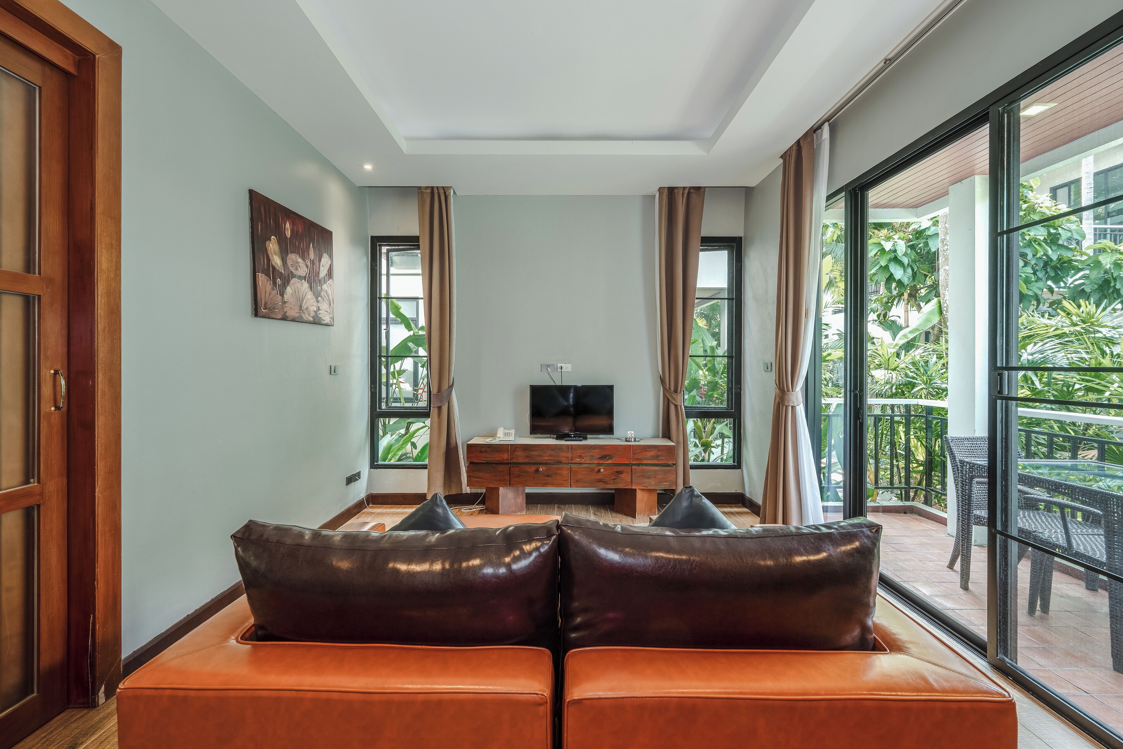 A living room with a sofa, a TV on a wooden cabinet, and large windows with curtains.
