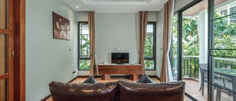 Una sala de estar con un sofá, un televisor sobre un mueble de madera y ventanas amplias con cortinas.